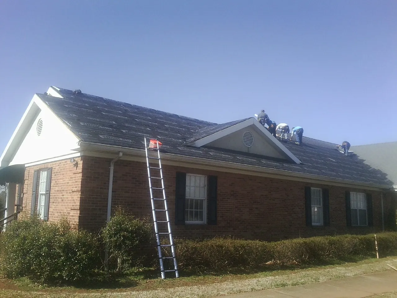Crew tearing shingles off a brick ranch roof with a ladder set — Metal Roof Repair work by our crew in New Lothrop