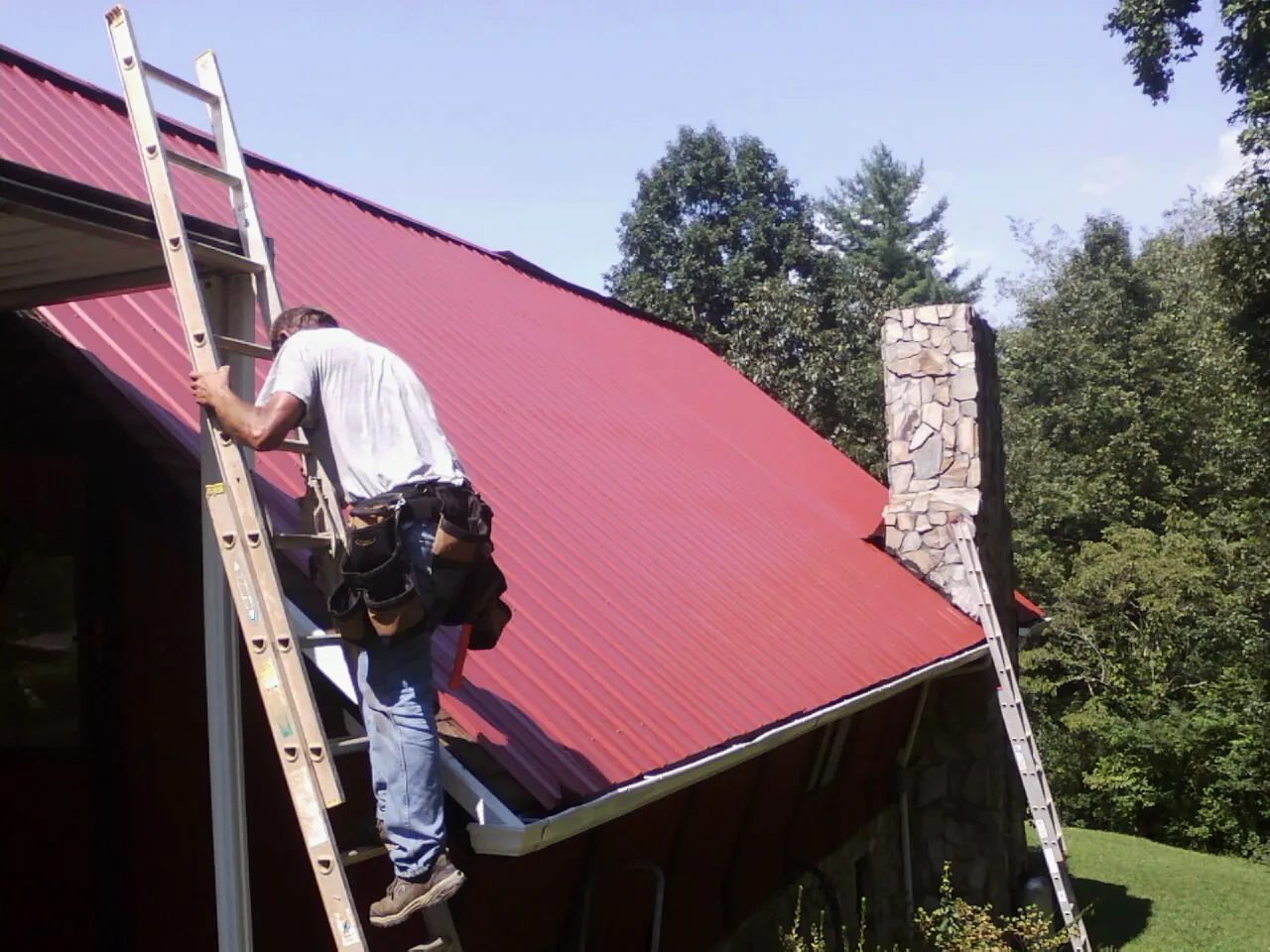 Roofer on a ladder beside a red metal roof and stone chimney — Soffit & Fascia Repair by Roofing Brothers in New Lothrop